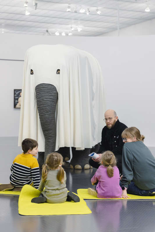 Des enfants assis face à un faux éléphant recouvert d'un drap.