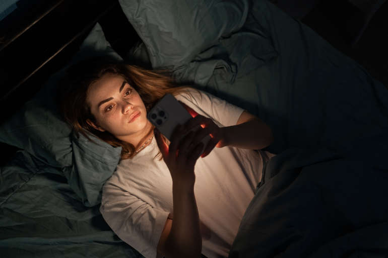 Une jeune femme à l'air fatigué regarde son téléphone dans son lit ©Adobe Stock